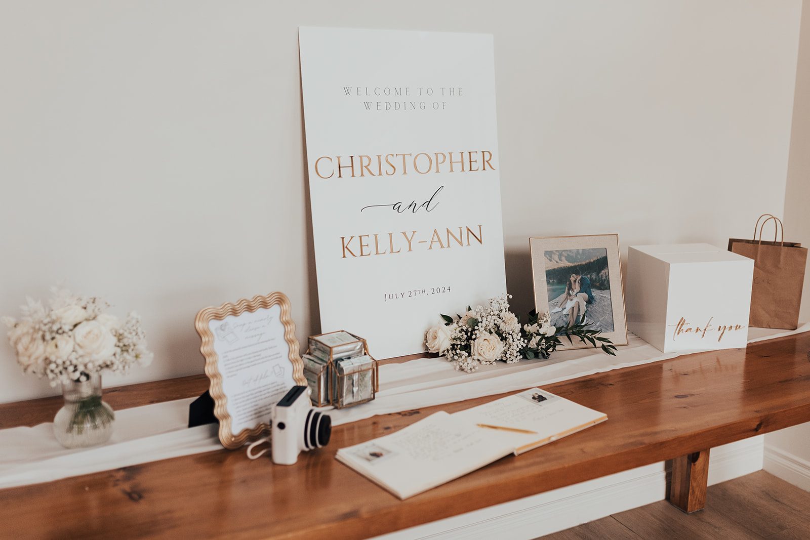 Mr + Mrs Day Wedding welcome table with a large white and gold sign for Christopher and Kelly-Anne, styled with framed photos, a guest book, candles, florals and a card box on a wooden bench.