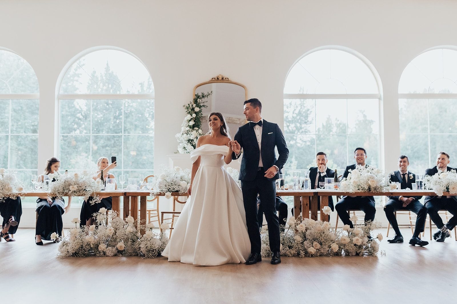 Mr + Mrs Day The bride and groom enter the reception holding hands and smiling, framed by their harvest style head table adorned with white floral arrangements and large arched windows behind them.