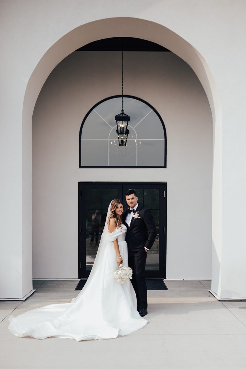 Happy Couple Bride and groom stand beneath a larch arched entrance, with the brides long train flowing behind her.