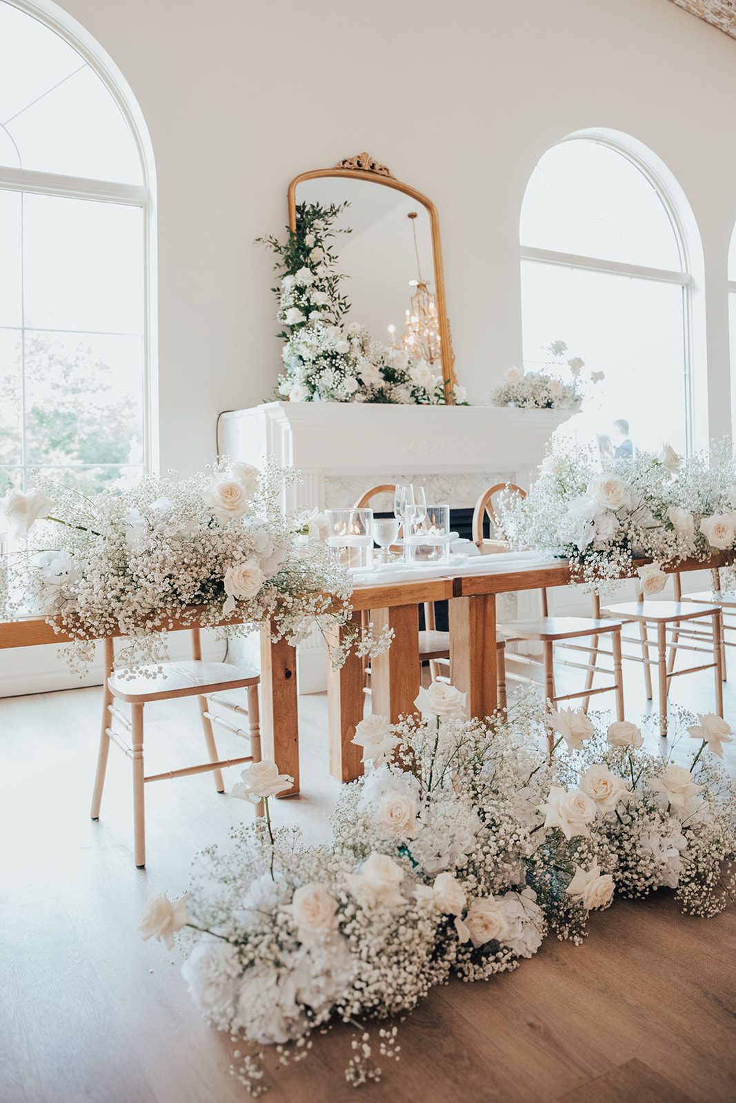 Mr + Mrs Day Head table decorated with fluffy white arrangements of baby's breath and roses in front of a fireplace with a gold mirror on the mantle, surrounded by natural light provided by the large arched windows.