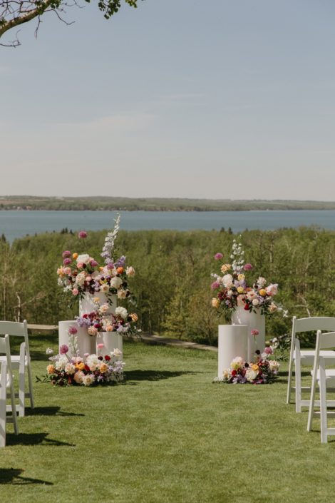 The Lakeview ceremony space at Hilltop Wedding Centre set up at their open house with three white pillars stacked on either side at the end of the aisle adorned in gorgeous arrangements of purples, peach, blues and burgundy instead of a floral arch.