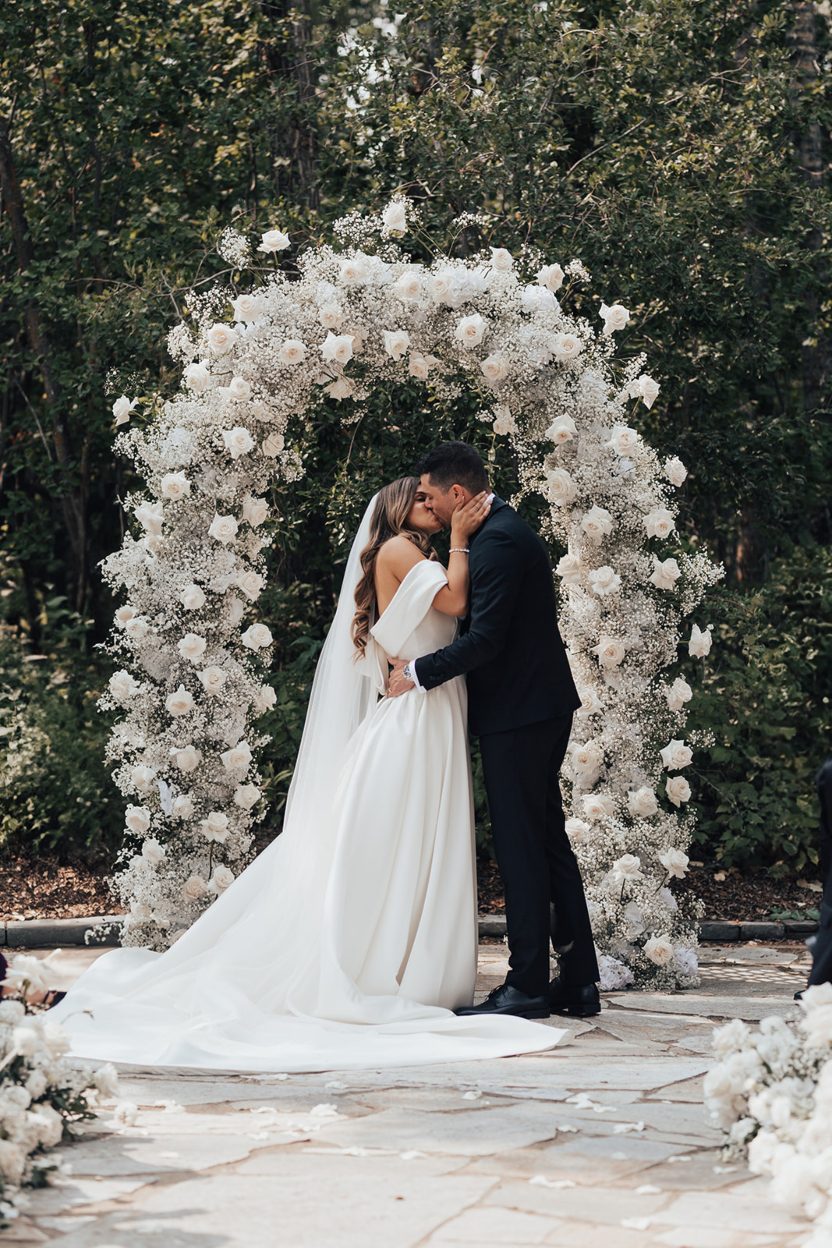 I do Bride and groom sharing their first kiss as husband and wife under a white rose and baby's breath arch surrounded by lush trees.