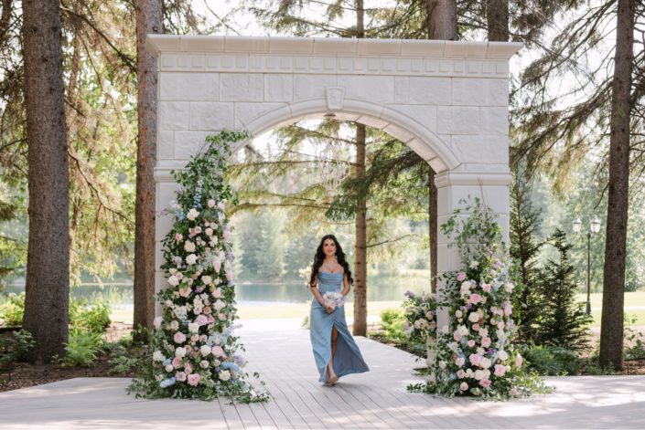 A bridesmaid in a gorgeous powder blue dress walks through the outdoor ceremony arch at Archway Manor, decorated with a broken arch in a mix of white, ivory, and mixed pinks with lush greenery.