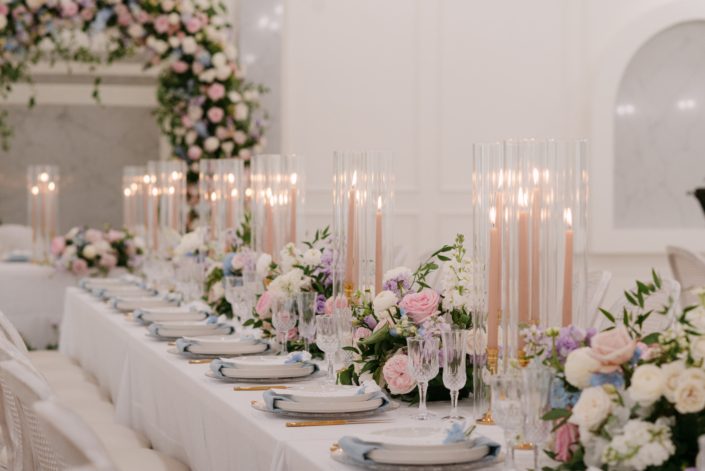 A magnificent wedding reception set up, a full floral arch in the background, with long rectangular guest tables set up with oodles of soft pink taper candles with lush arrangements of pinks, blues and ivory in-between.