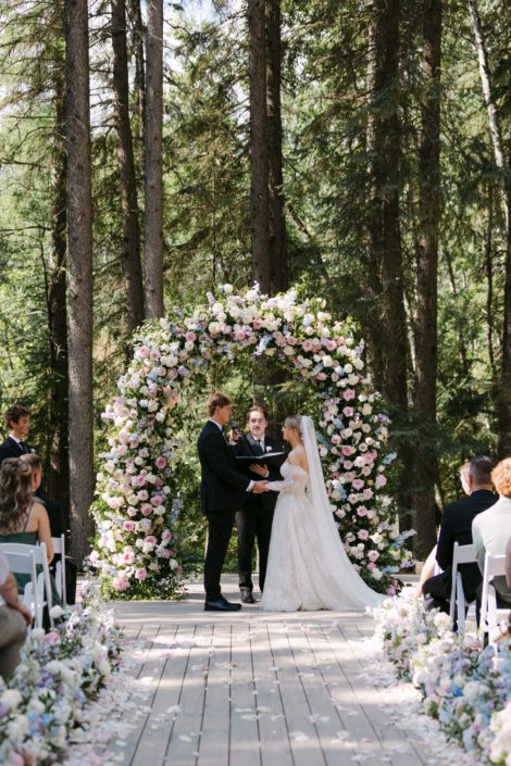 A show topping full floral arch at the outdoor ceremony space at Archway Manor, featuring an assortment of pinks and whites and accented with lush greens. A Calyx Couple stands in front, hands clasped mid ceremony.