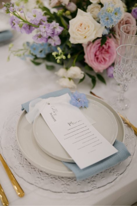 A close up of a place setting showing a menu, accented with an individual delphinium bloom, blue table linens, crystal glassware, gold flatware and part of the gorgeous centrepiece featuring huge pink garden roses, ivory spray roses, blue delphinium and other stunning gardeny blooms.