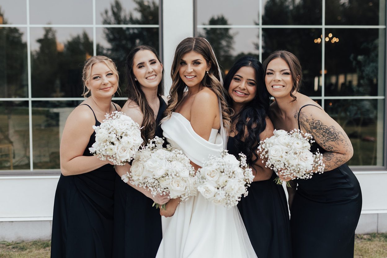 Kelly Ann + Bridesmaids Bride with her bridesmaids standing close together in front of arched windows holding matching white bouquets.