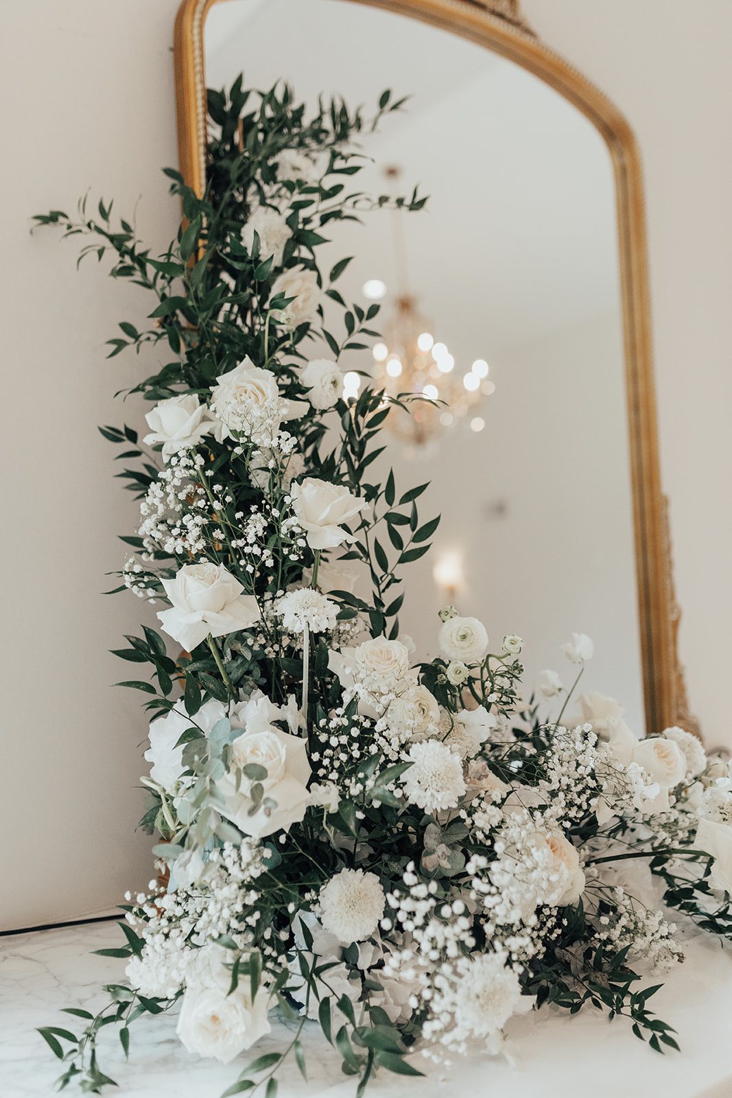Mr + Mrs Day Detailed view of lush mantle arrangement featuring white rose, ranunculus, baby's breath and greenery climbing up a gold mirror.