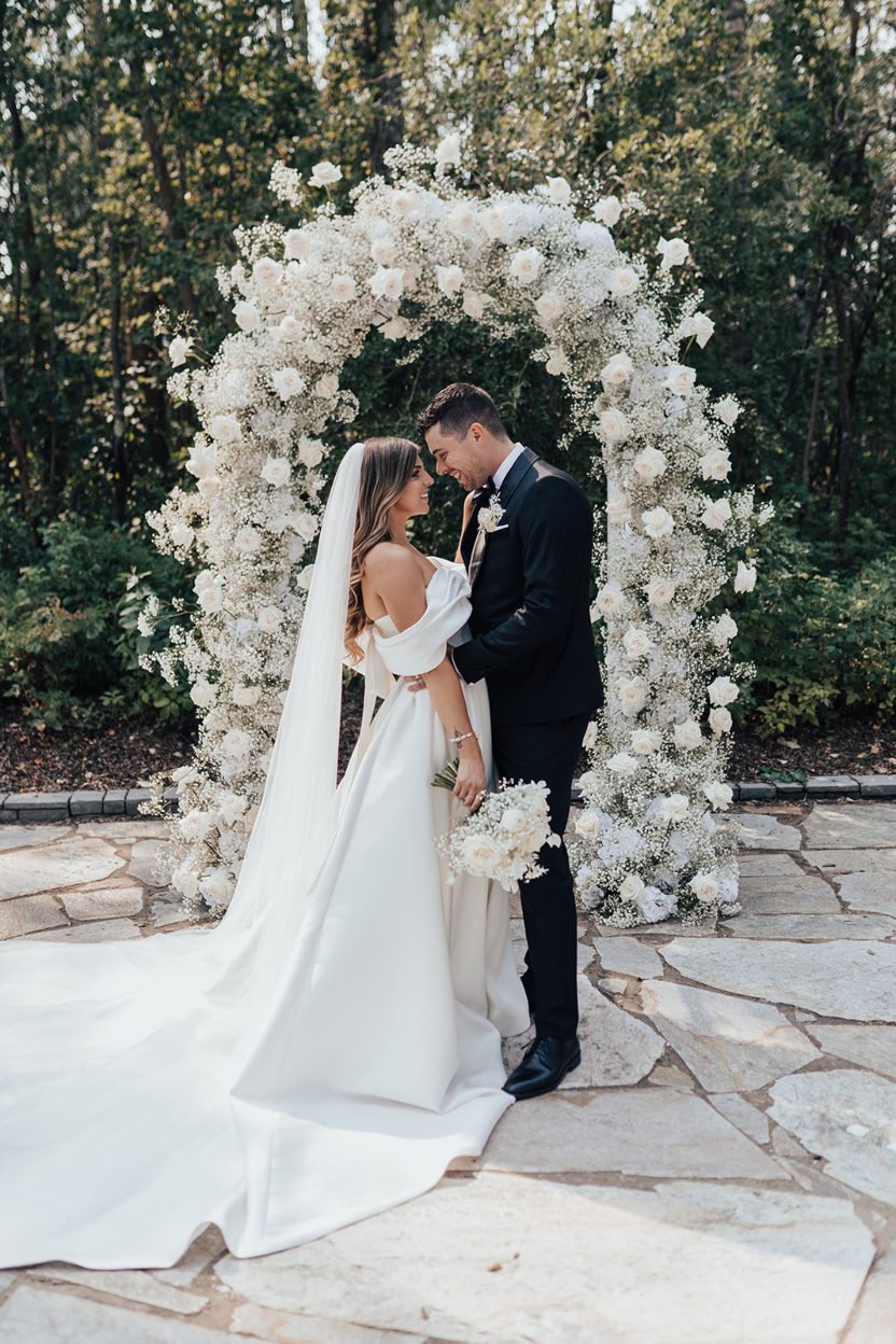 Mr. + Mrs. Day Bride and groom standing together with their foreheads almost touching in front of a lush floral arch of white roses and baby's breath.