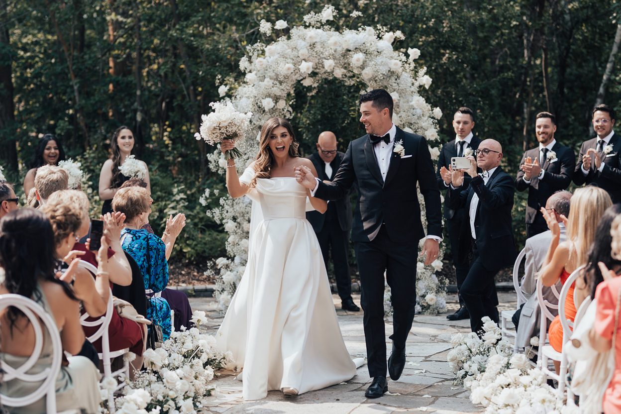 Mr. + Mrs. Bride and groom walking back up the aisle smiling as guests cheer, with lush white floral arch behind them.