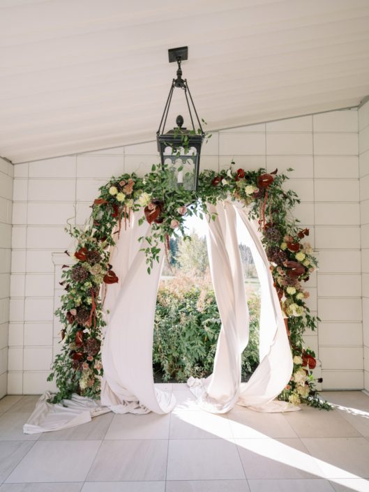 Outdoor space at Archway Manor decorated for a Professionals Lunch with tons of lush, organic feeling greens paired with rich burgundy displayed around one of the arches along with soft billowing fabric which is blowing softly in the breeze in the picture.