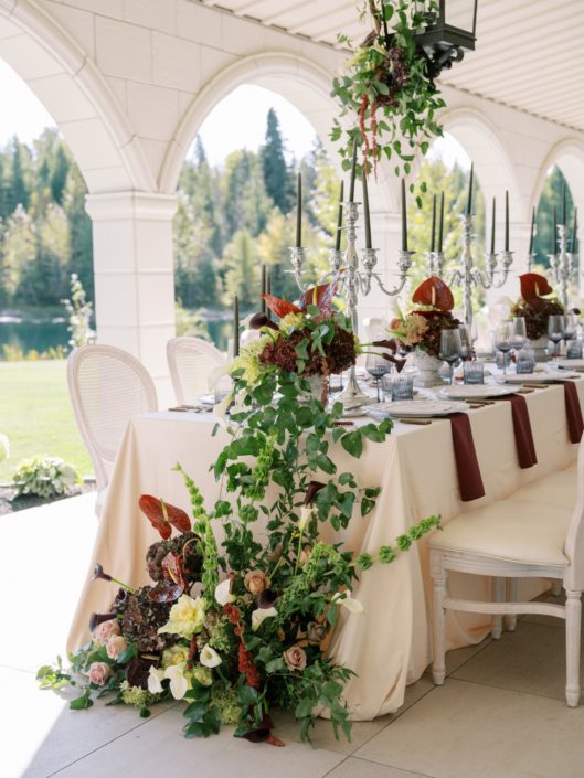 The outdoor space at Archway Manor set up for a Professionals Lunch, a big arrangement in burgundy and green displayed in front of the table on the floor, along with several centrepieces down the table and even flowers hanging from a lantern above the table.