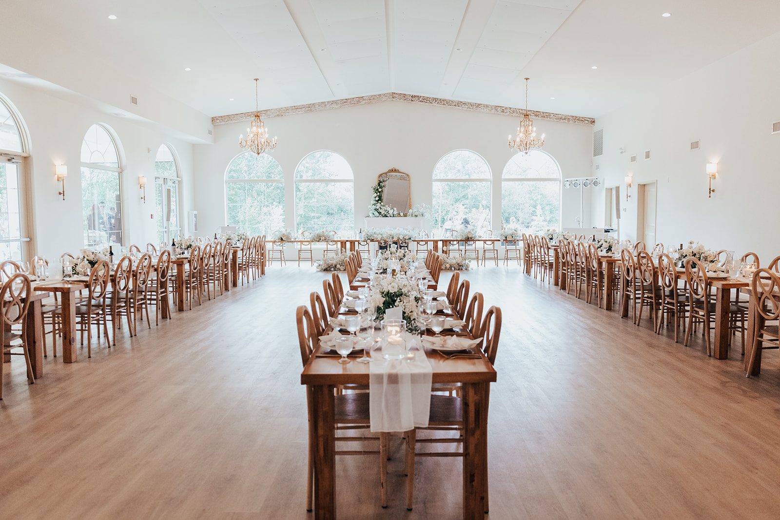 Mr + Mrs Day Wide ceremony style view of the bright reception hall with long wooden harvest tables, white florals, arched windows and chandeliers at Sparrow Lane.
