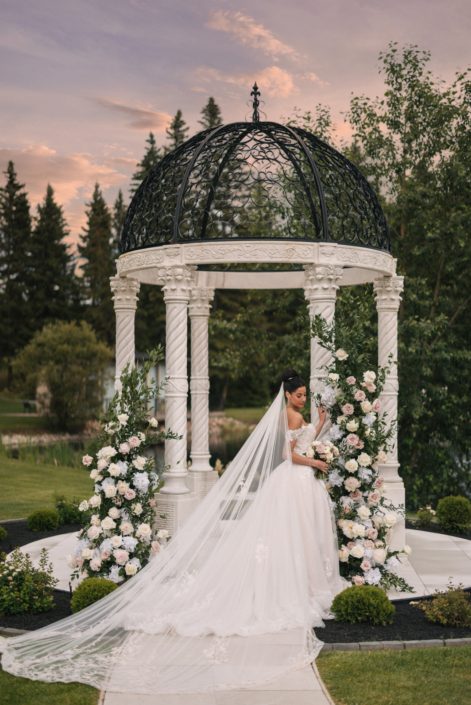 Our Calyx bride stands at the gazebo at Archway Manor which has been decorated with a broken arch design in whites and blush blooms as the sky is an array of pretty pastel shades at sunset.