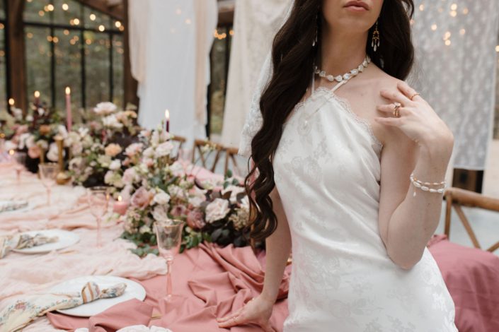 Our model bride in the "True to Hue" photoshoot stands in front of a table, with her hand at her shoulder showing off a beautiful pearl necklace. Behind her a stunning table with blush table runner, big centrepieces of blush, neutrals and burgundy with pretty vintage fabrics hanging behind the table.