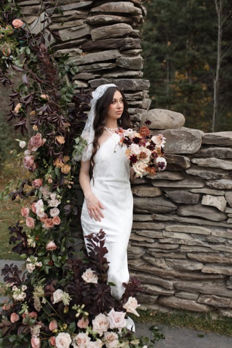 Our model bride in the "True to Hue" photoshoot holds a stunning bouquet in mixed neutrals, blush and burgundy while standing next to the lush floral arch.