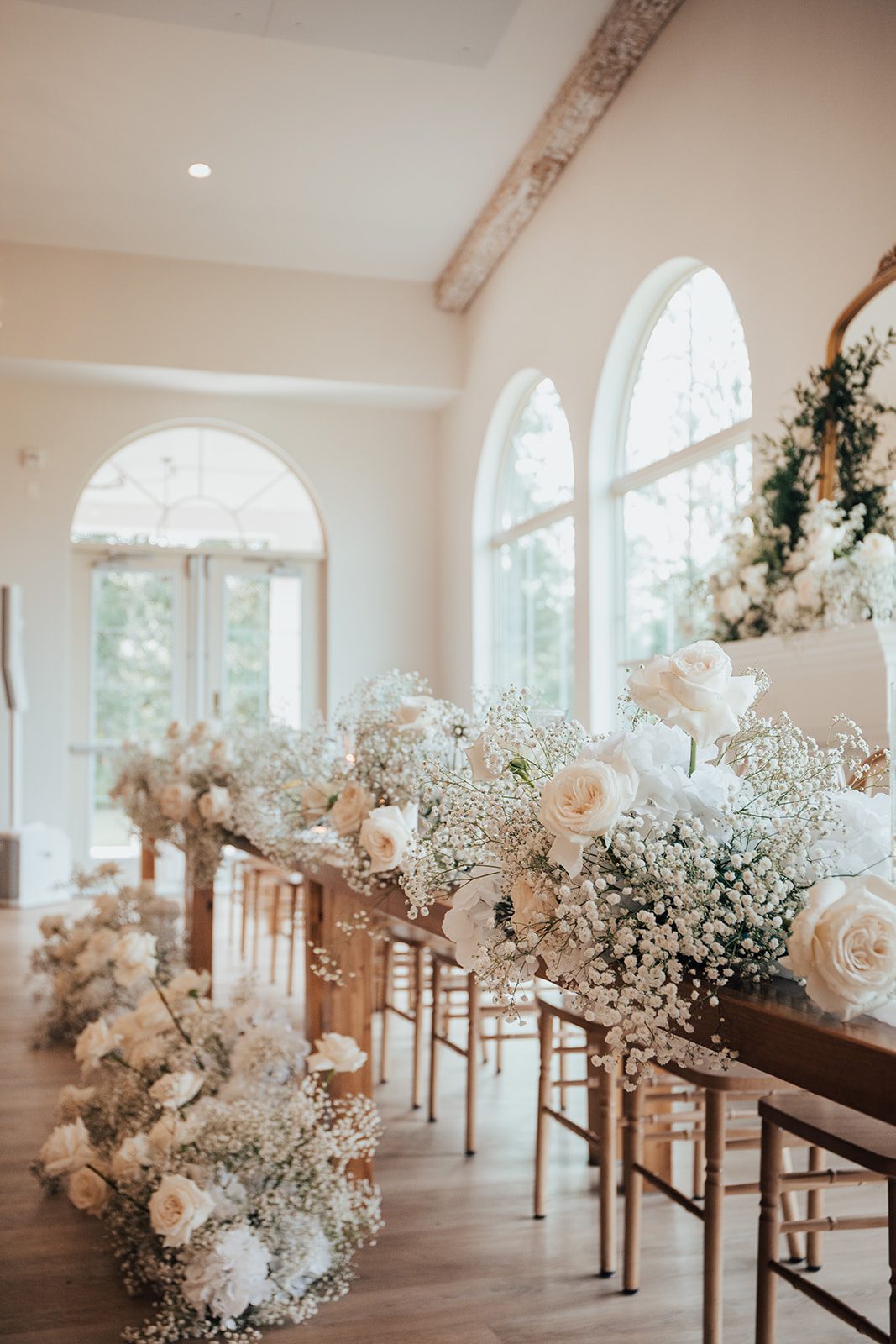 Mr + Mrs Day Row of long wooden tables for the head table lined with clouds of white roses and baby's breath, illuminated by natural light from the arched windows.