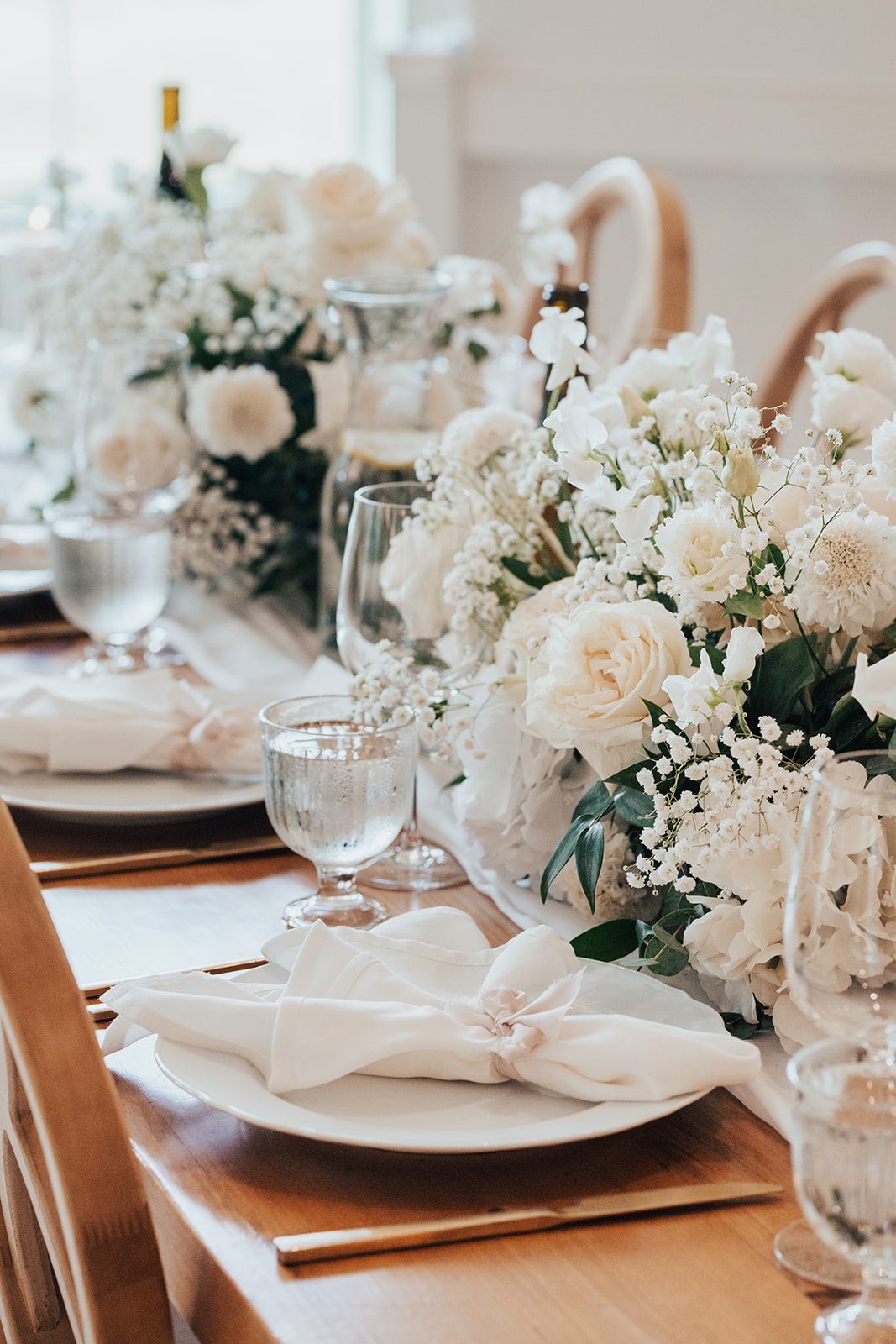 Mr + Mrs Day Close up of white floral arrangements running down the length of the reception tables featuring roses, ranunculus, and baby's breath paired with white linens and plates and accent with gold flatware.