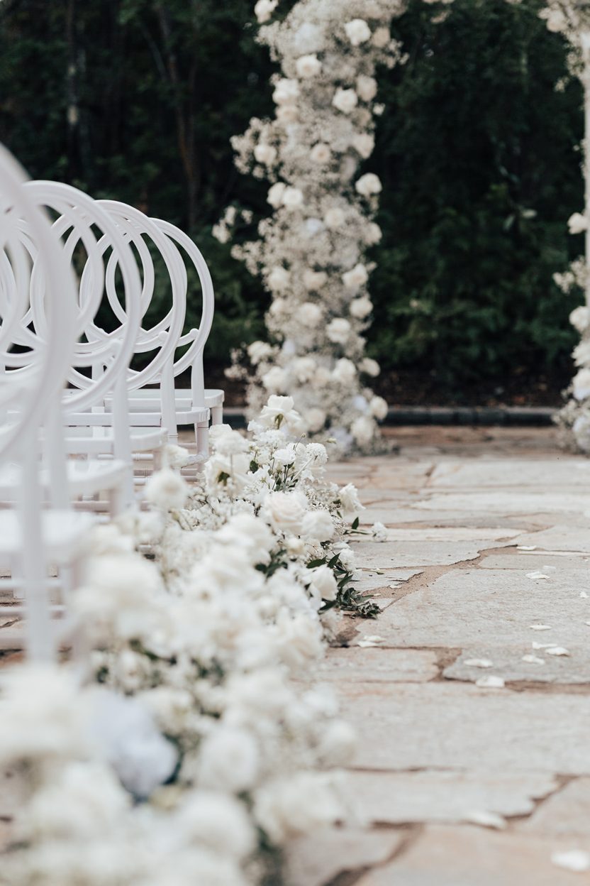 White Florals Side view of white ceremony chairs and low floral arrangements lining the stone aisle leading to the floral arch.