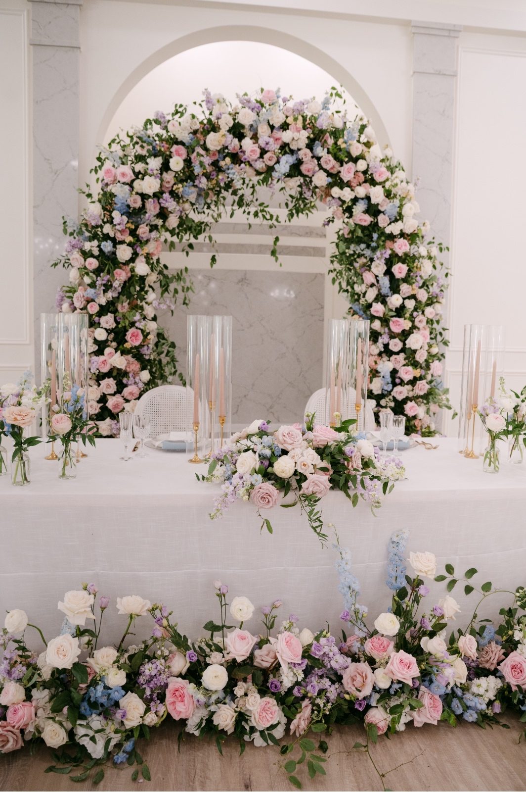 Kaylee and Oran's sweetheart table with the amazing, full floral arch behind the table, bud vases and blush tapered candles on the table, and gorgeous pastel arrangements on the floor in front of the table for a cozy and romantic space for the couple to share during dinner.
