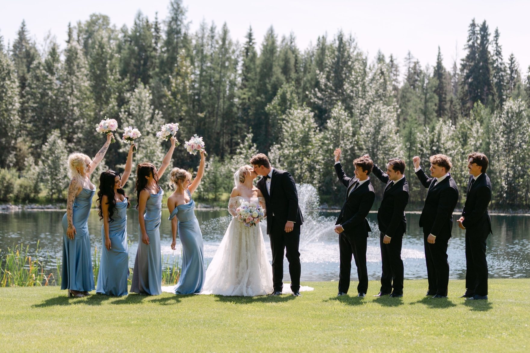 Oran and Kaylee with their wedding party in front of the pond, the bridesmaids to the left and the groomsmen to the right. Oran and Kaylee share a kiss and the wedding party have their hands up in the air, celebrating.