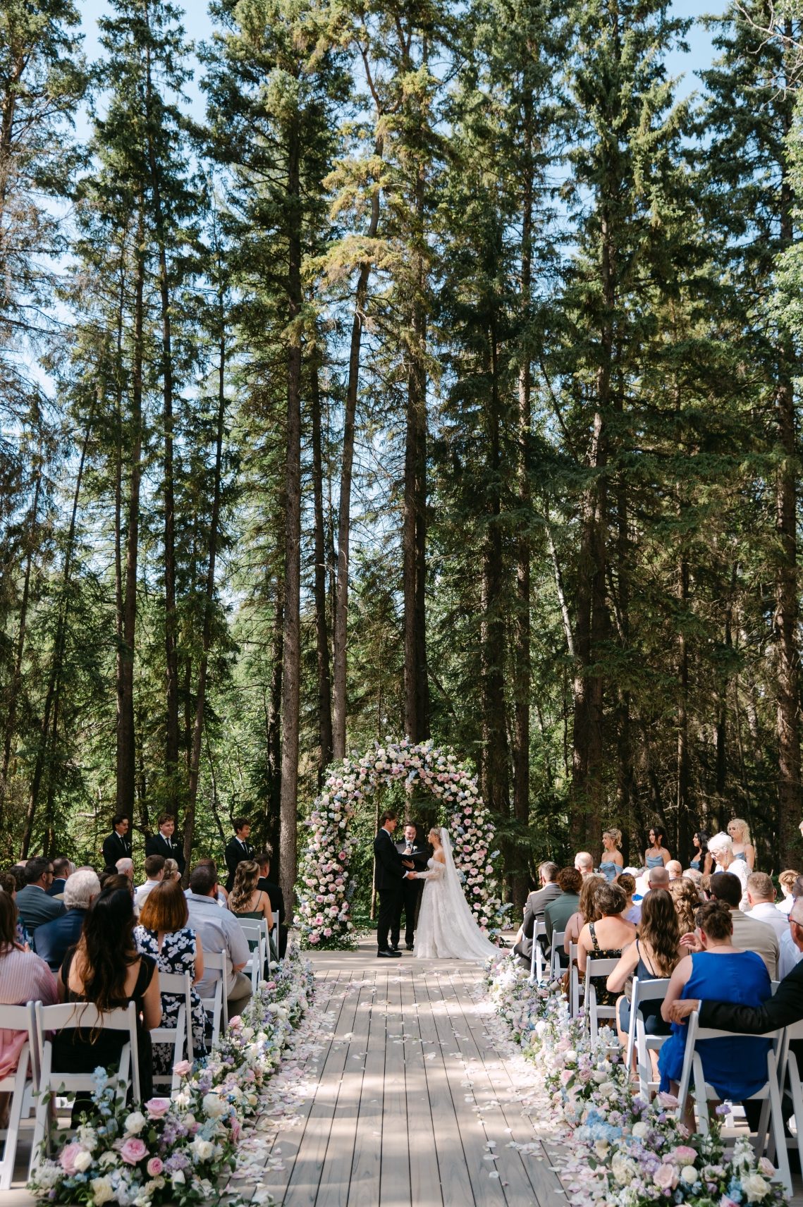 The ceremony has begun, Oran and Kaylee are standing in front of the floral arch, with the huge forest trees in the background.