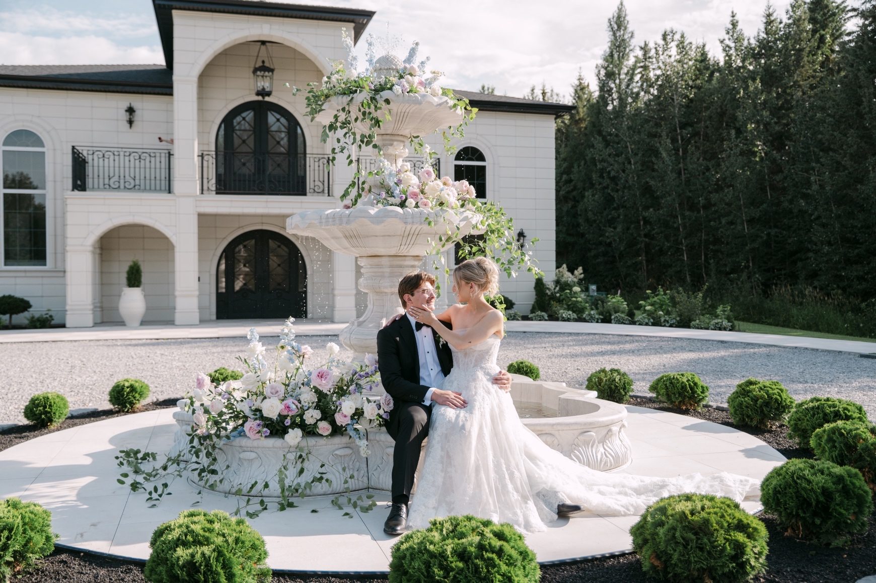 Oran is sitting on the edge of the fountain and Kaylee is sitting on his lap and they are gazing lovingly at each other, with the stunning view of Archway Manor in the background.