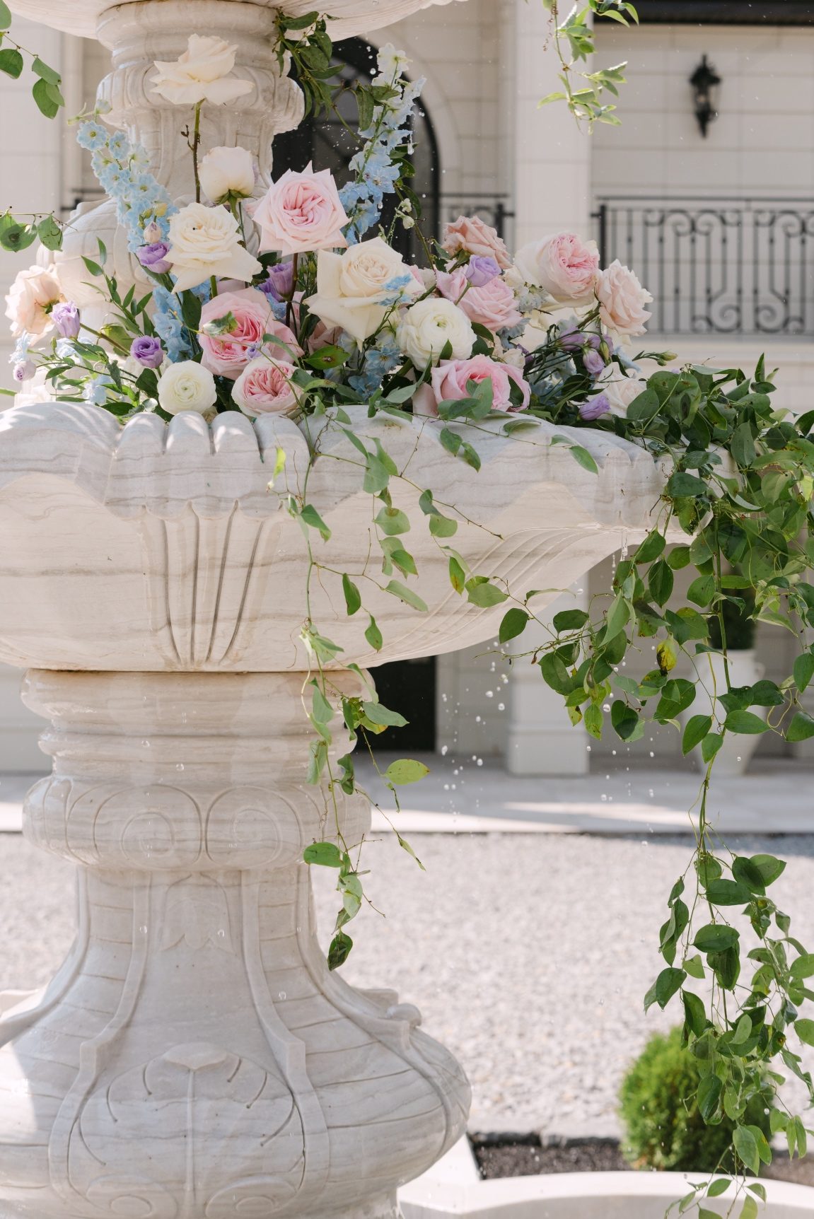 A close up of the second tier of the fountain with a big, beautiful arrangement of roses, delphinium, ranunculus, stock and delphinium, with foliage pooling over the edge.