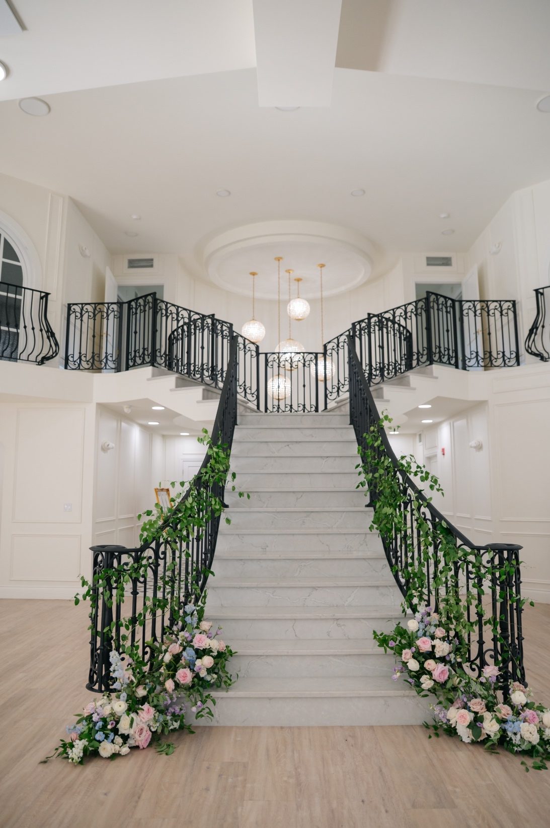 The grand staircase at Archway Manor with large pastel floral arrangements at the base, with foliage woven up the railings and banister in an organic fashion.