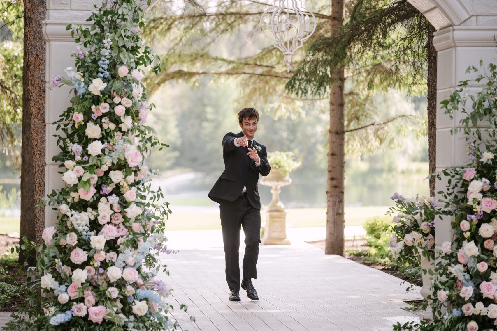 In front of the stone arch, Calyx designed a stunning broken arch, overflowing with roses, delphinium, ranunculus lisianthus and greenery. One of the groomsmen is headed in, being a bit cheeky and pointing at the camera as he walks in.