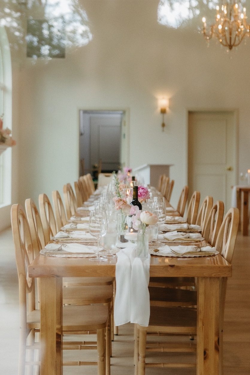Guest Tables The view down the length of the long harvest table for the guests with the chairs neatly lined up, soft linen draping over the edge of the table and bud vases of pastel blooms placed down the length of the table.