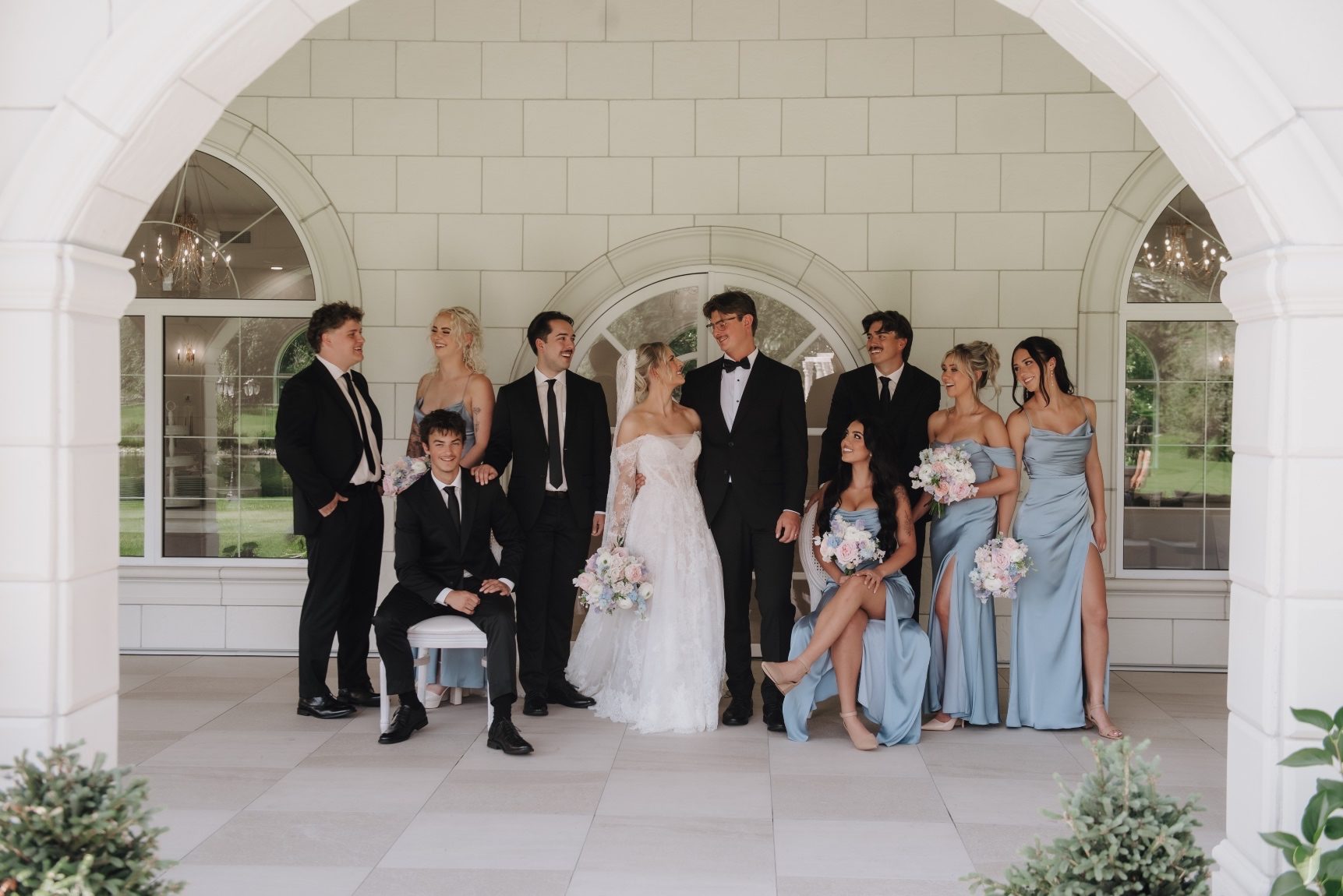 Oran and Kaylee stand in the centre looking at each other, while their bridal party (some sitting, some standing) surrounds them, looking at them)