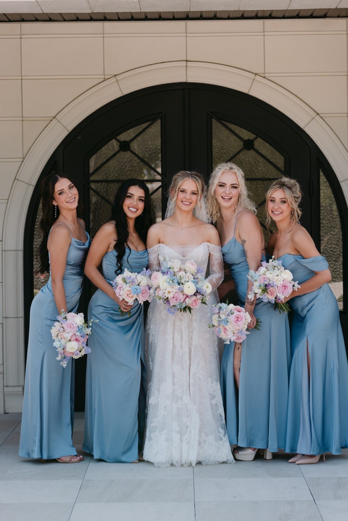 Kaylee and her bridesmaids stand in front of one of the arches at the venue, holding their bouquets and smiling at the camera.
