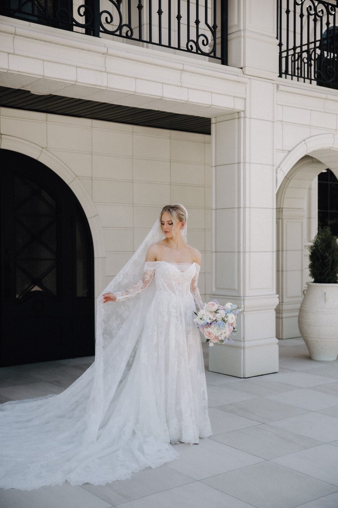 Kaylee stands outside Archway Manor, she is holding her bouquet in one hand and touching the edge of her veil with her other hand, while it fans out past her feet.