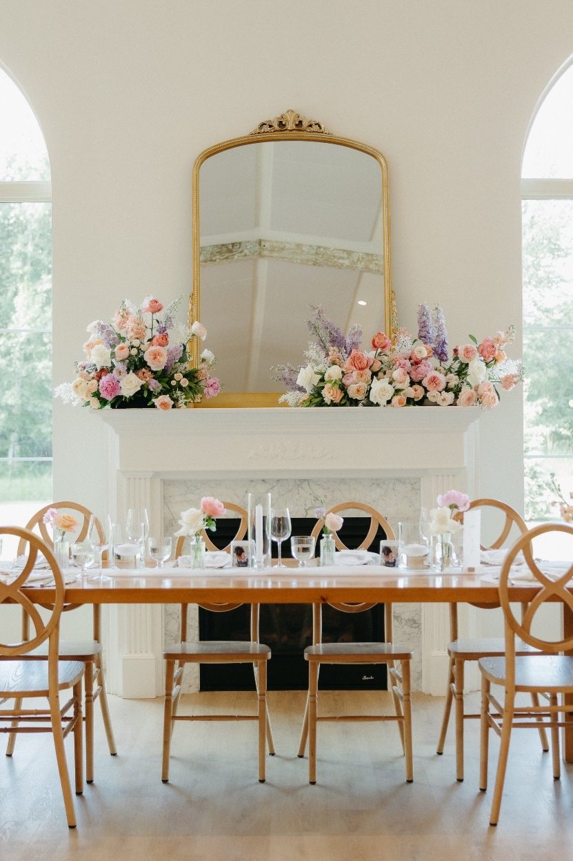 Stunning Head Table Elegant head table display with simple bud vases of pastel blooms, in behind a fireplace with a mantel with two large pastel arrangements and a beautiful gold framed mirror.
