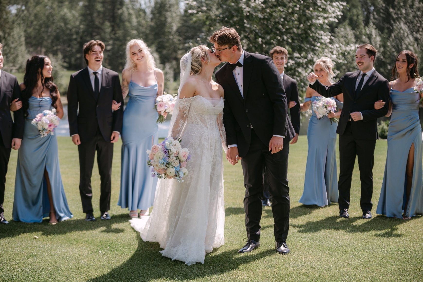 Kaylee and Oran share a kiss with their wedding party slightly behind them split off in pairs.
