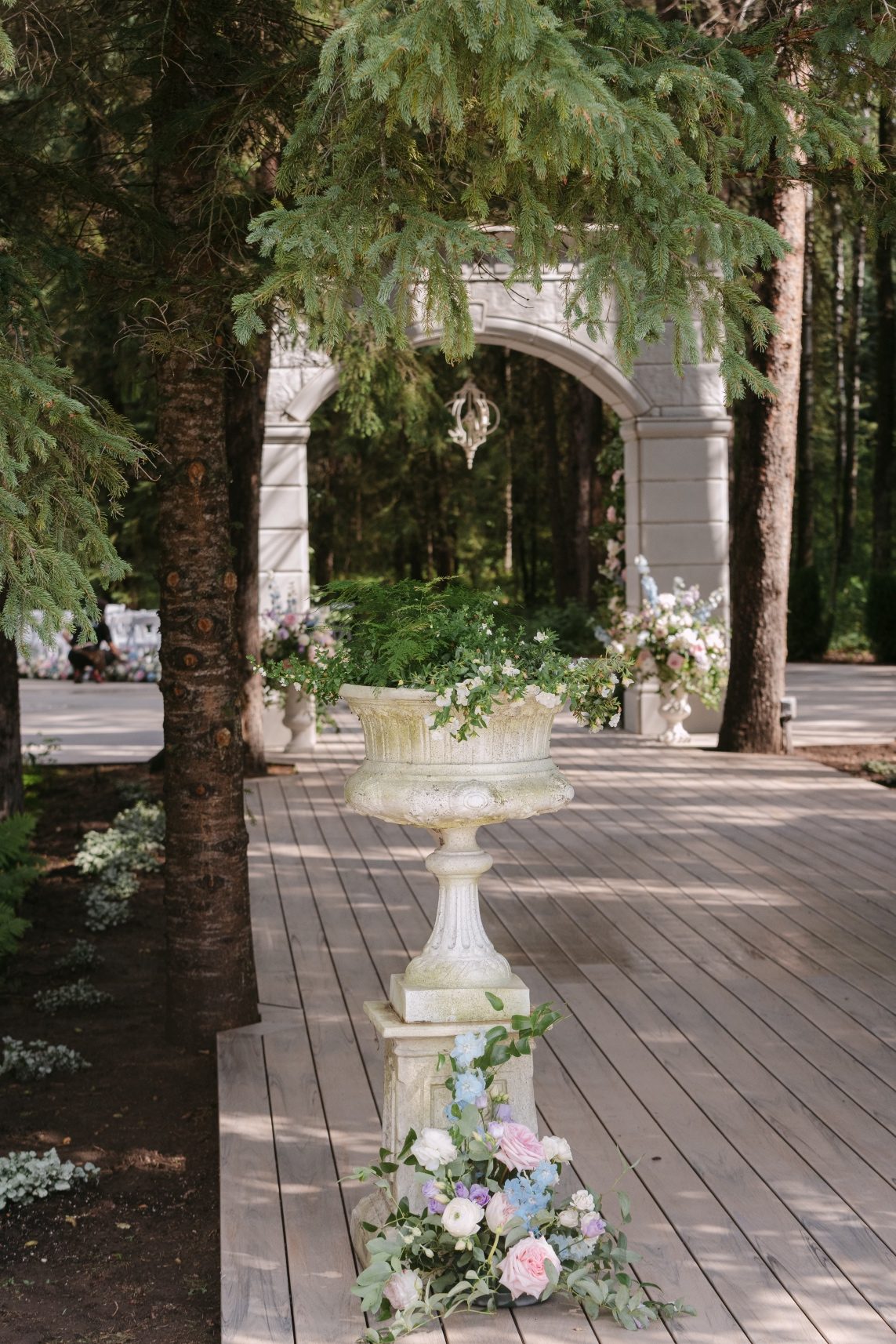 Leading into the ceremony space, we are greeted by an ornate planter, with pastel arrangement at its base and in the background the stone arch with two large floral urn arrangements.