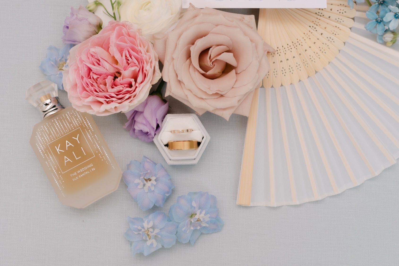 A close up of the rings in the flatlay image surrounded by blue delphinium blooms, a pink garden rose, a soft blush rose, and purple lisianthus blooms.