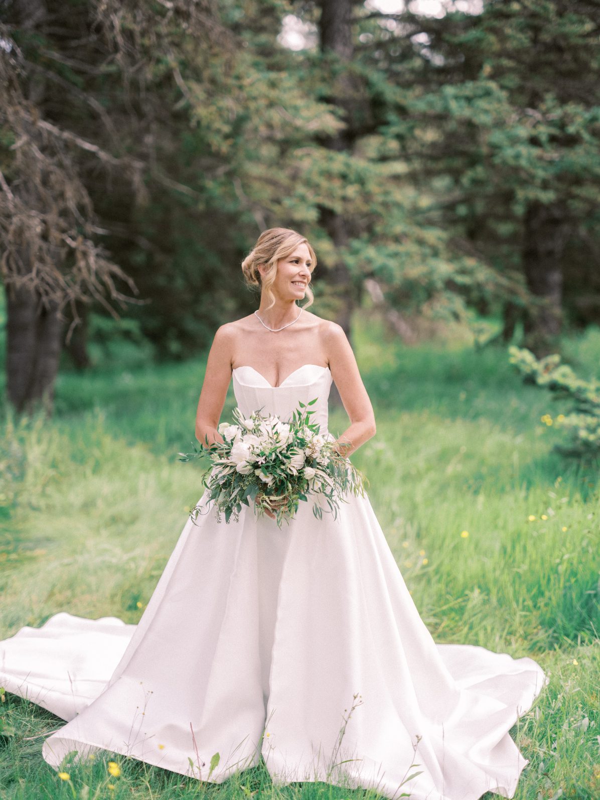 Blushing Bride Bride in strapless satin wedding gown holding a lush white and green bouquet in a forest meadow.