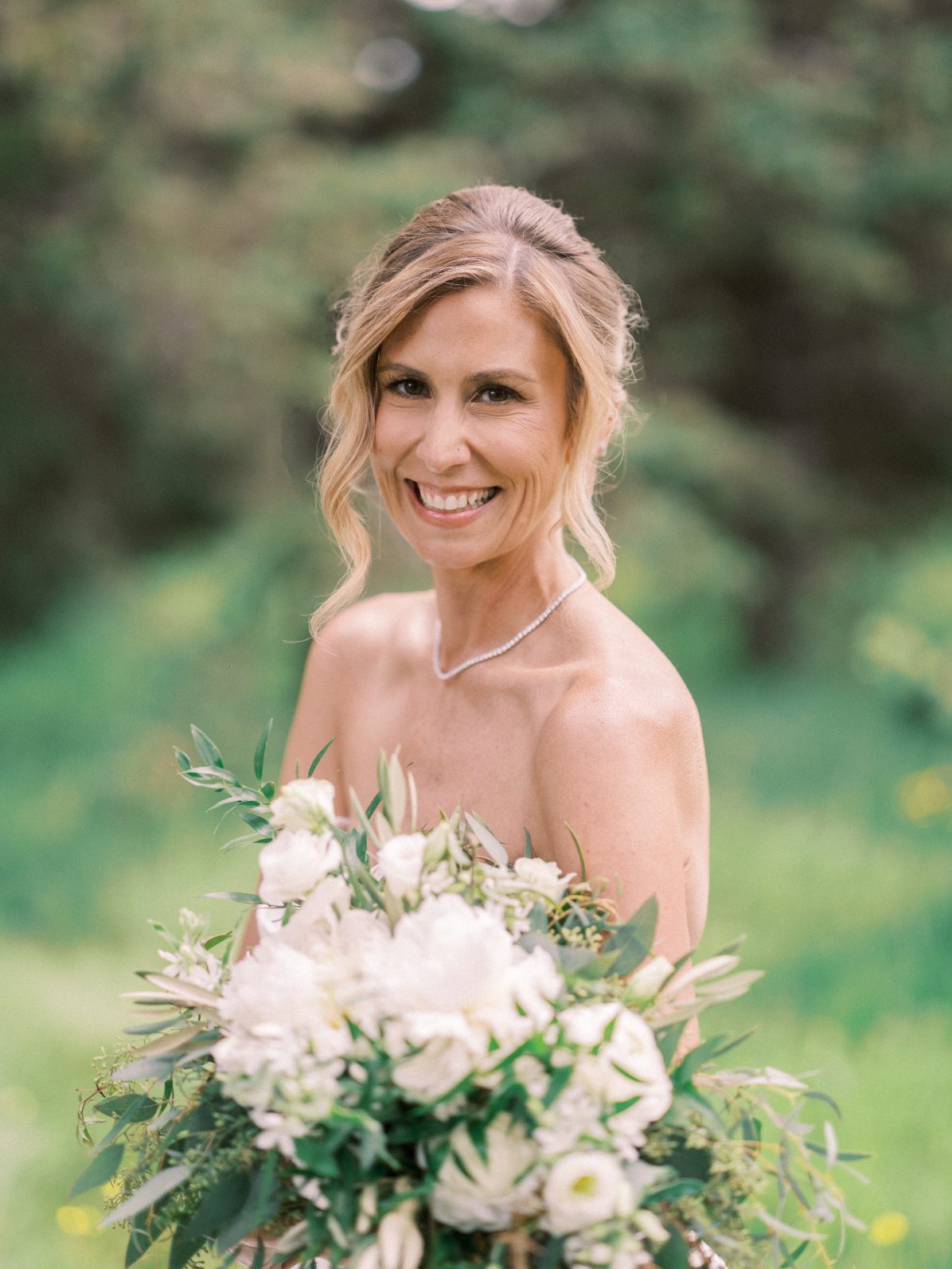 Calyx Bride Portrait of bride smiling while holding white and greenery bouquet in a natural forest setting.