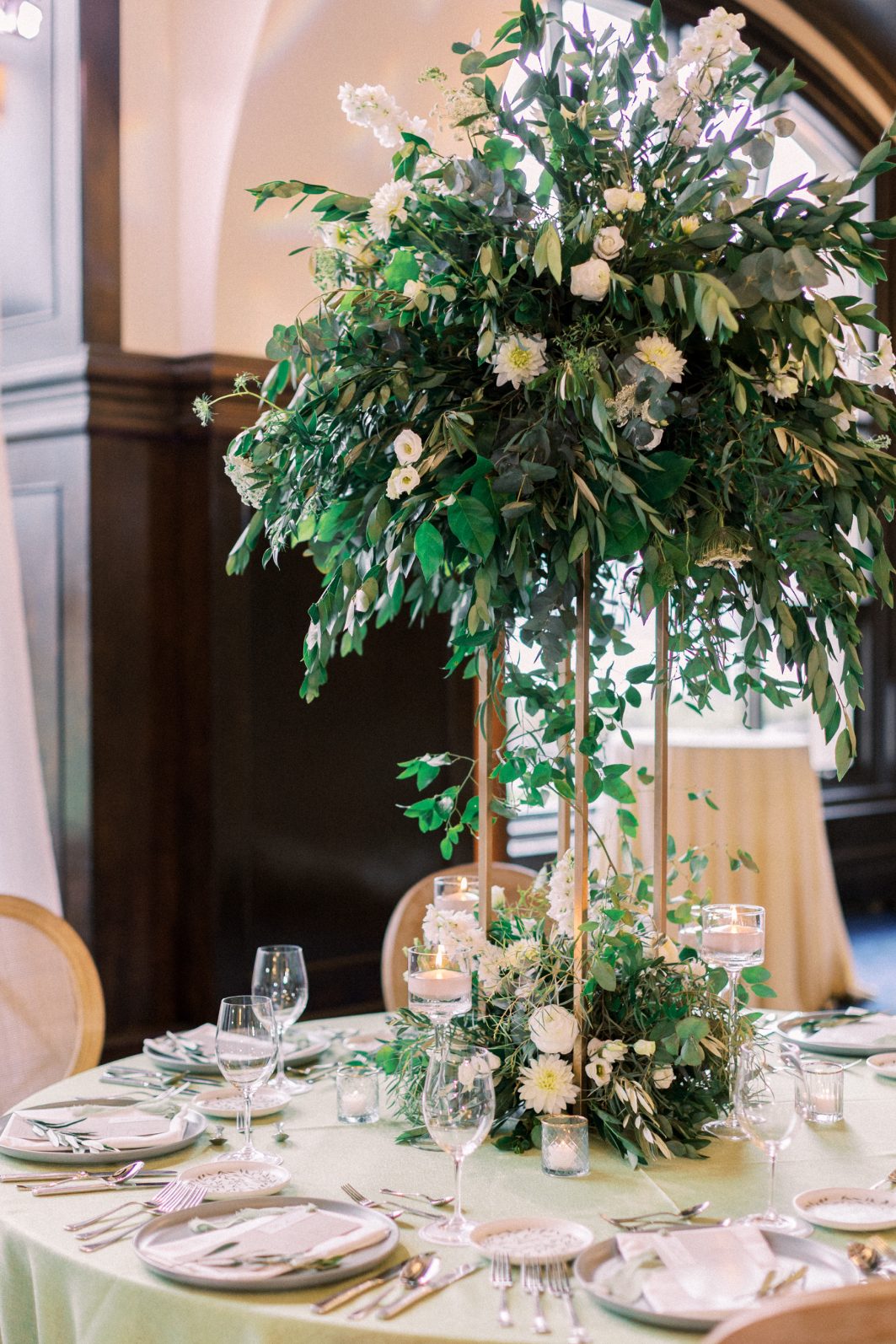Calyx Centrepiece Tall reception centrepiece overflowing with greenery and white blooms above candlelit round table.