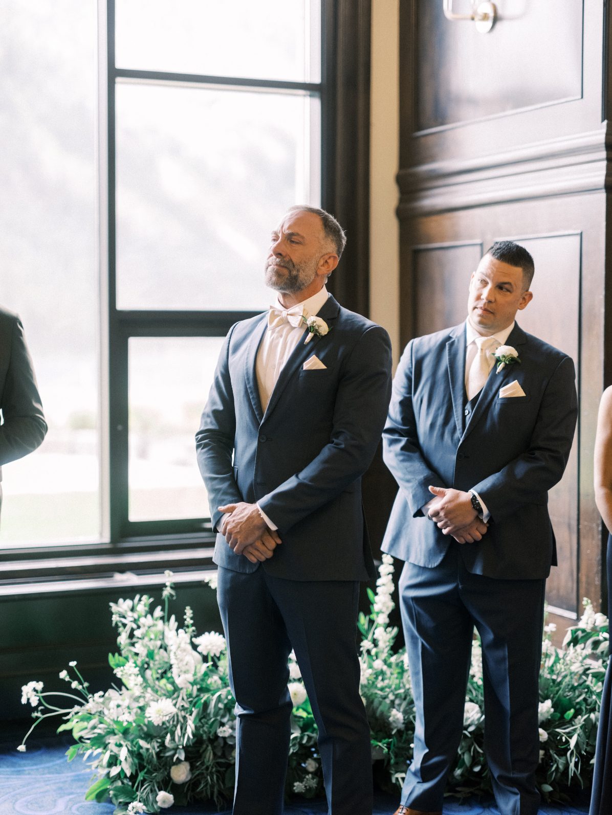 Calyx Groom Groom and groomsmen standing beside abundant white ceremony florals framed by grand arched windows.