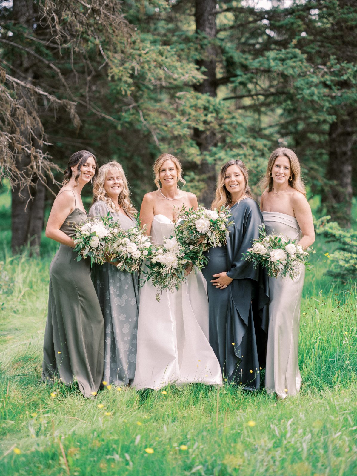 Candace + Bridesmaids Bride with bridesmaids in coordinated green and neutral toned dresses holding organic white bouquets outdoors.