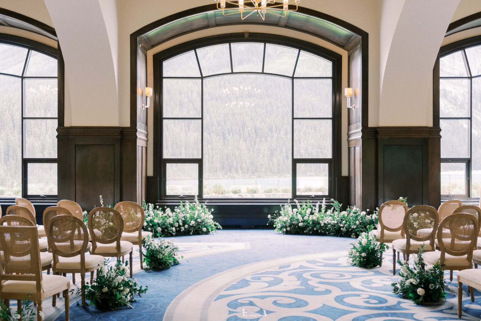 Ceremony Setup Elegant indoor ceremony space featuring arched windows, can back chairs, and low white floral installations along the aisle.
