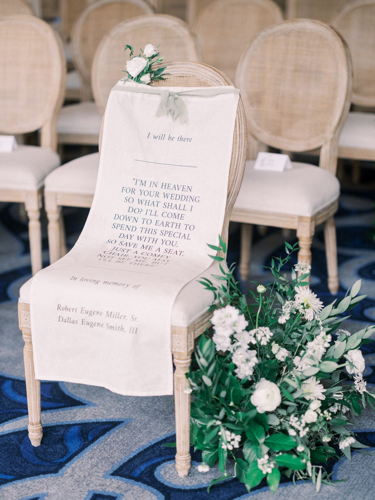 Ceremony Tribute Ceremony chair adorned with a memorial sign and delicate white floral arrangement accented with lush greenery.