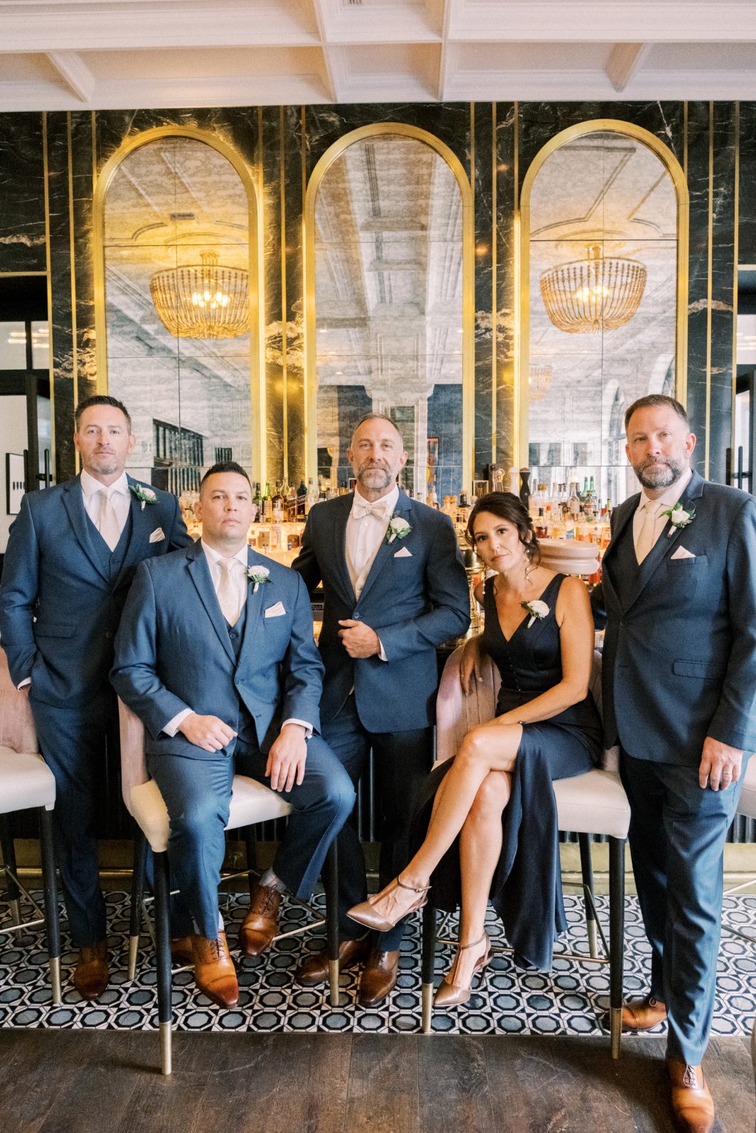 Groom + Groomsmen Groom and groomsmen in blue suits with white boutonnières posing inside a stylish bar.