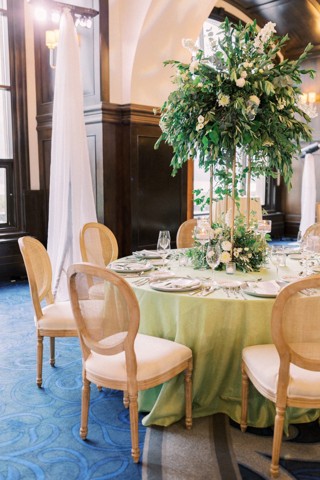 Guest Tables Round reception table with towering greenery centrepieces, candlelight accents, and when cane back chairs.