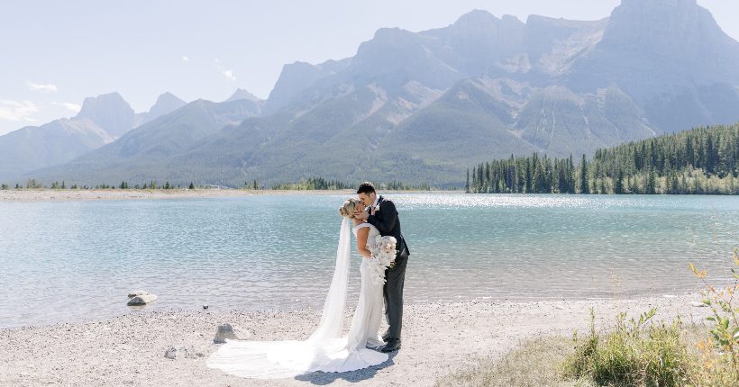 H&J Feature Thin Helen and James share a kiss in front of beautiful blue waters of a lake, with mountains in the background.