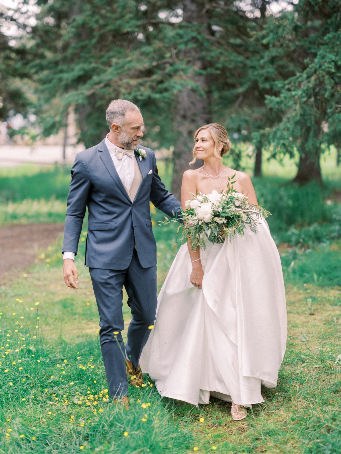 Happy Couple Bride and groom walking through a lush meadow, the bride carrying an organic white and greenery bouquet beneath towering evergreens.
