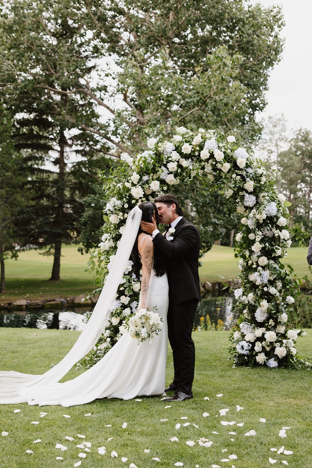 Luxury Wedding ceremony at Hilltop wedding center featuring a full round top archway with smilax and greenery accented by white flowers, white roses as well as a bride being dipped by the groom holding a bride bouquet